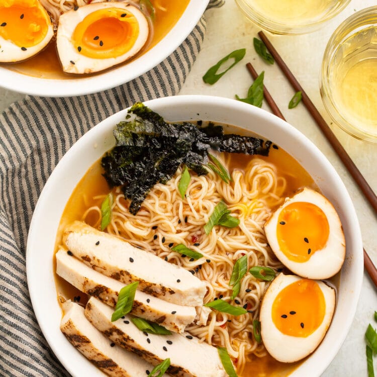 Top-down view of two white bowls holding spicy ramen noodles in broth with slices of chicken, two yolky egg halves, and green garnish.