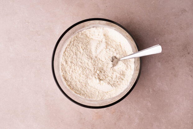 Overhead view of flour, baking soda, and salt whisked together with a fork in a large glass mixing bowl on a pink surface.