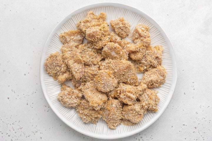 Breaded boneless, skinless chicken breast chunks on a large white plate ahead of being skewered.