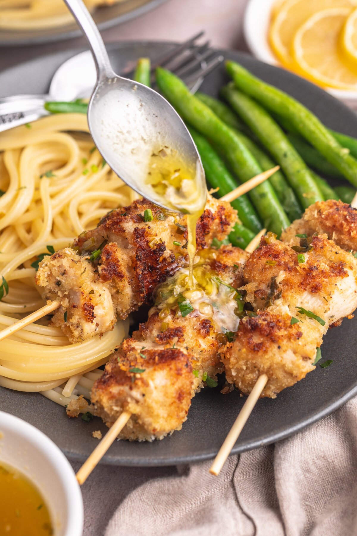 Herb butter sauce being drizzled over three chicken spiedini skewers plated with angel hair pasta and bright green beans.