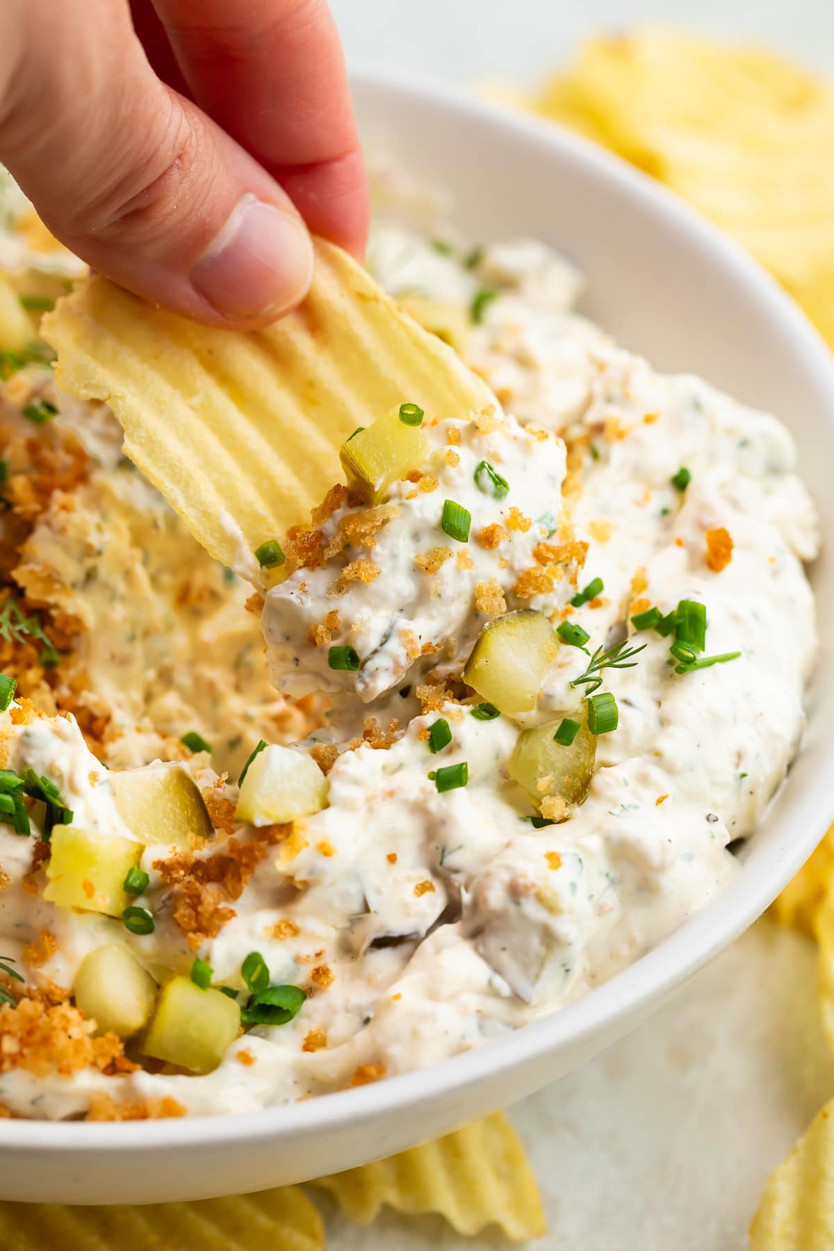 A white woman's hand holding a potato chip with ridges, scooping fried pickle dip out of a bowl.