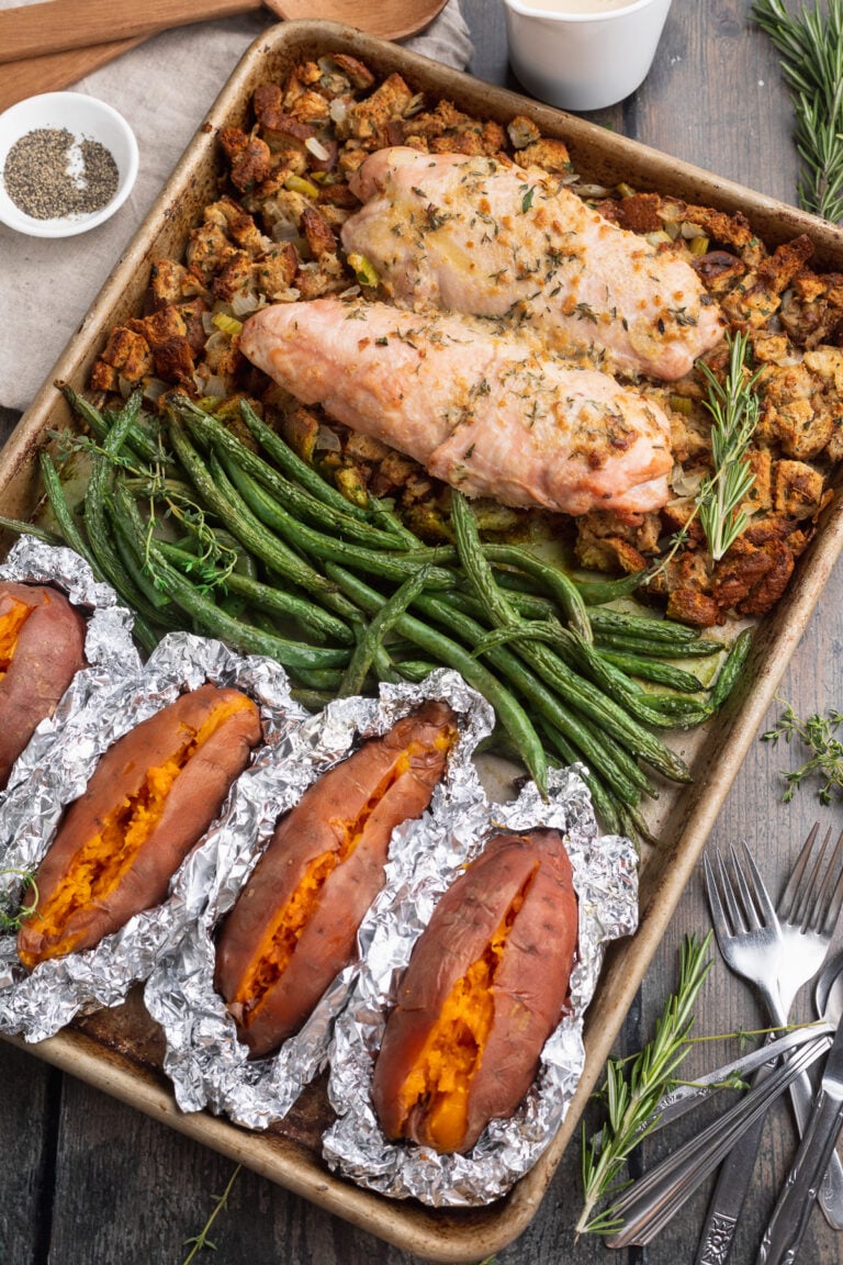 Overhead view of a sheet pan, resting at an angle, filled with green beans, turkey, foil-wrapped sweet potatoes, and stuffing.