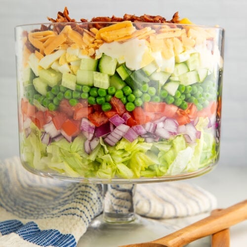 A 7 layer salad in a glass trifle dish with a pedestal base next to wooden utensils and a dish towel.