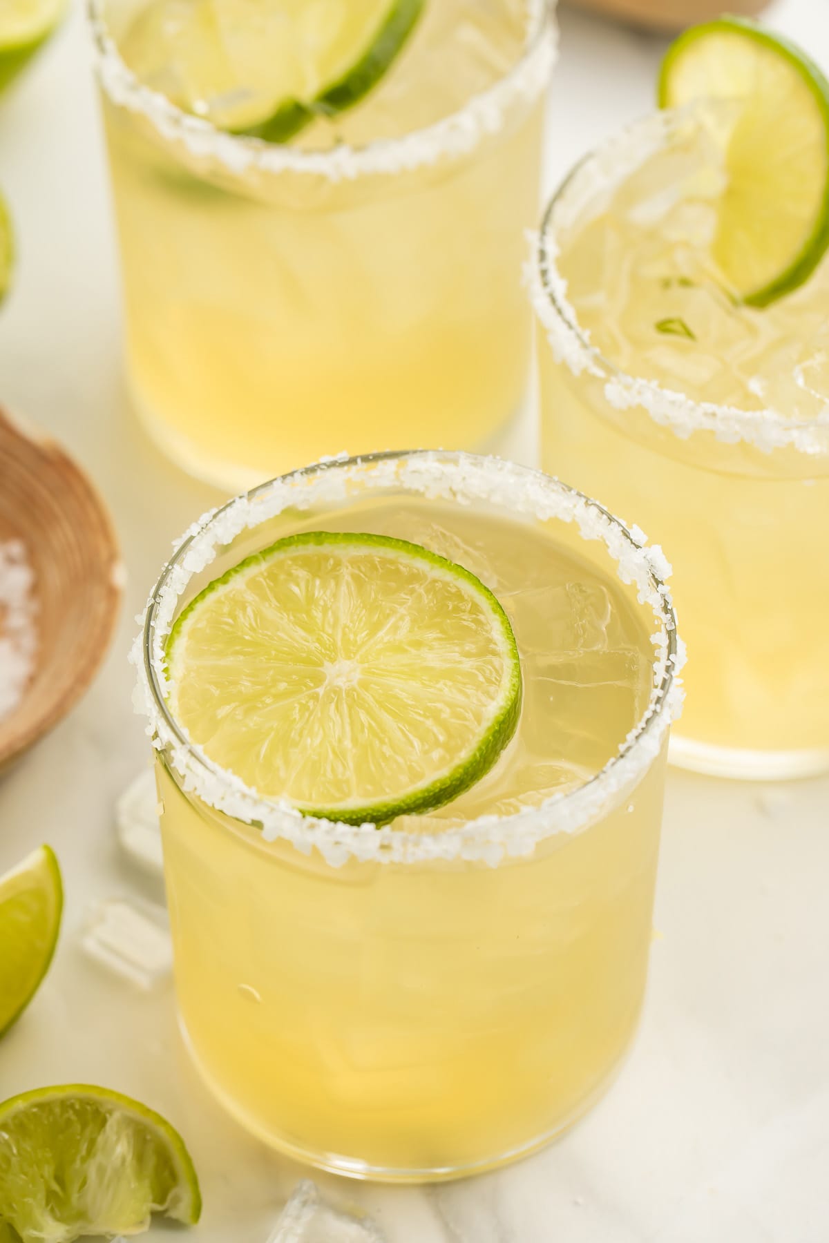 A salt-rimmed glass of virgin margarita garnished with a lime coin in front of 2 other glasses of margaritas.